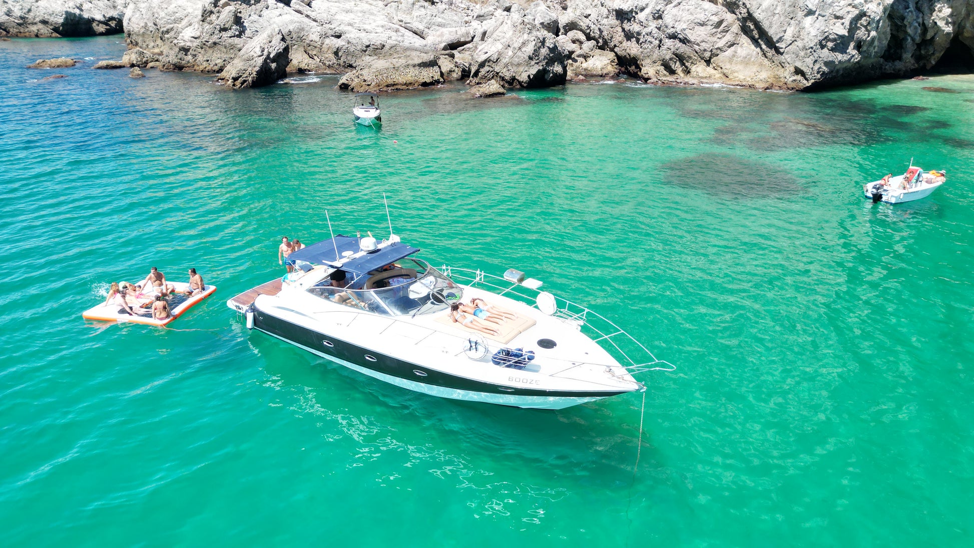 Luxury motor boat anchored in crystal clear emerald cove with floating platform and guests, limestone cliffs backdrop, Arrabida Natural Park, Setubal Peninsula, The Untold Weekend
