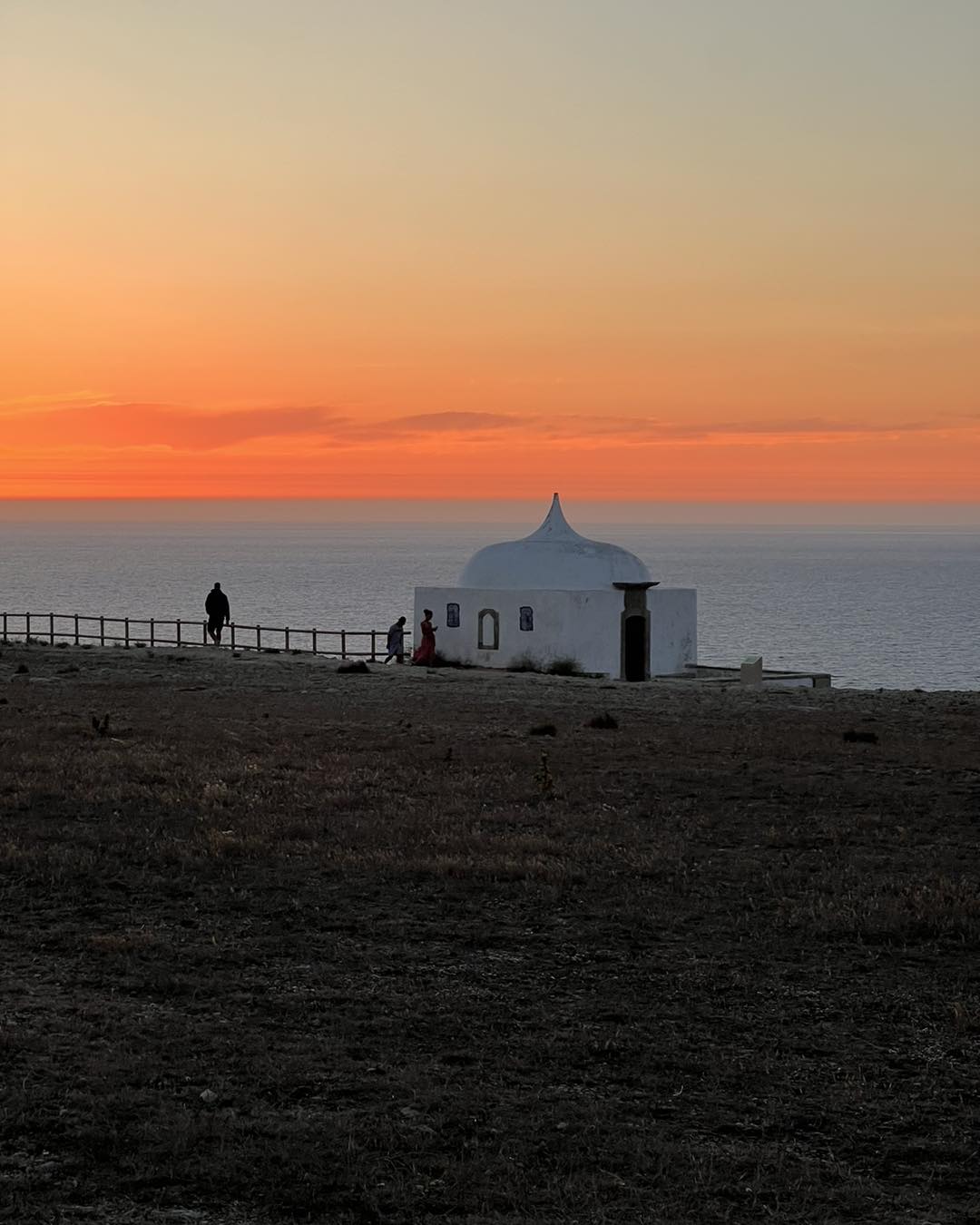 Dramatic orange sunset over Atlantic ocean with white ermida chapel silhouette and visitors at Cabo Espichel, Setubal Peninsula, Segredos da Arrabida Portugal